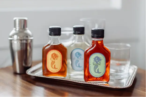 Small bottles of The Shipwright's Daughter cocktails on a silver tray with a cocktail shaker and glassware.