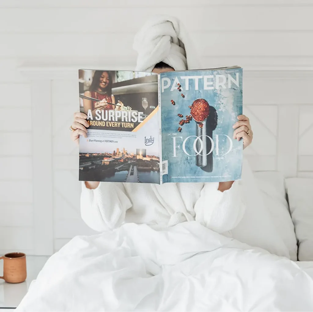 A woman in a white bathrobe and head towel relaxing in bed while reading a Pattern food magazine.