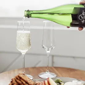 Sparkling wine being poured from a green glass bottle into a flute glass on a table with a snack board.