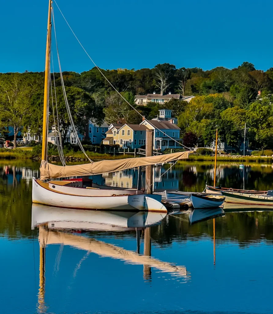 Sailboats anchored in a calm harbor with coastal houses and autumn foliage reflected in the blue water.