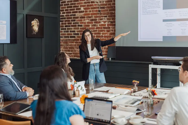 A businesswoman gives a professional presentation to colleagues in a modern, brick-walled office meeting room.