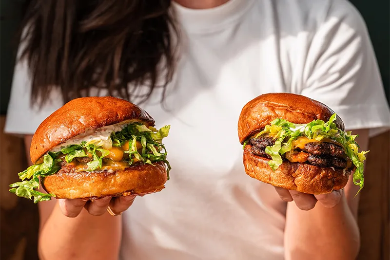 Person holding two delicious cheeseburgers with lettuce and toasted brioche buns.