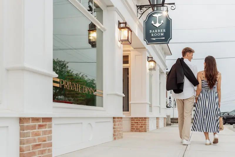 A couple walks past a white storefront featuring a Barber Room sign and private dining signage. They are looking for Things To Do in Mystic CT.