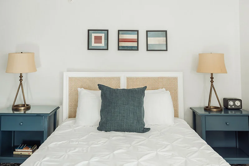 Modern bedroom with a white bed, cane headboard, teal accent pillow, and matching navy blue nightstands with lamps.