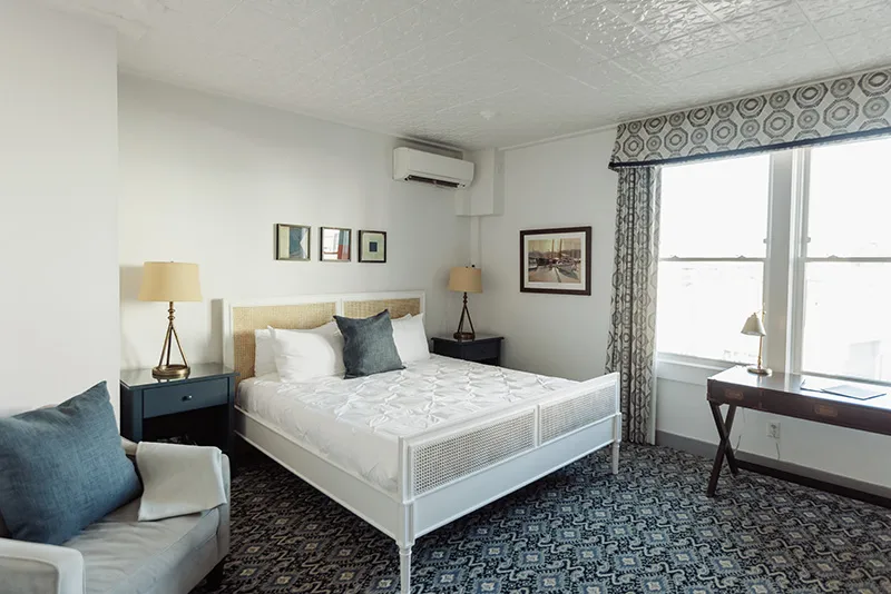Bright and cozy hotel bedroom featuring a white rattan bed, patterned carpet, and a sunny window seating area.
