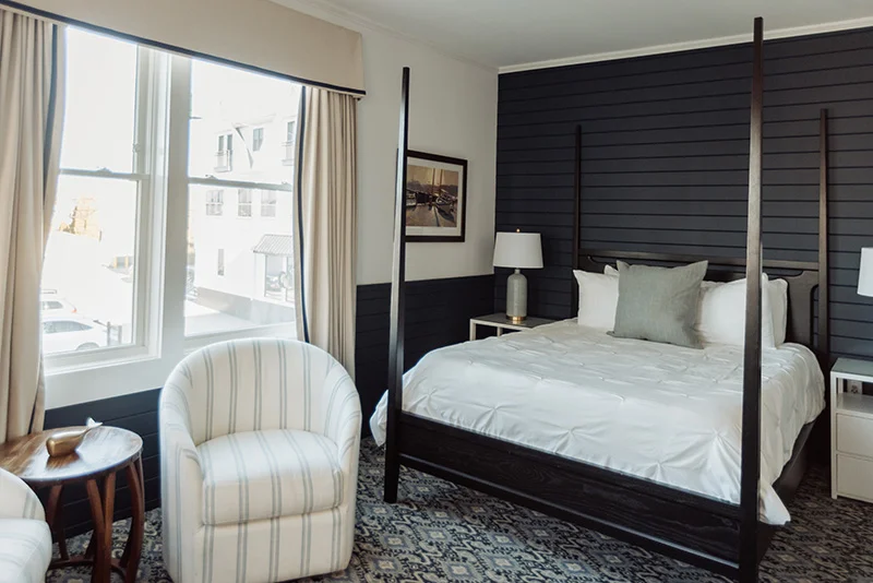 Modern hotel bedroom featuring a dark wood canopy bed, striped accent chair, and large windows with neutral curtains.