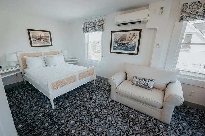Bright, coastal-style hotel bedroom featuring a white rattan bed, patterned carpeting, and a cozy armchair.