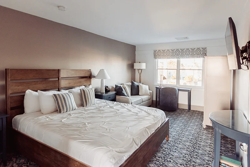Cozy hotel bedroom featuring a queen bed, seating area, and a functional workspace with patterned carpeting.