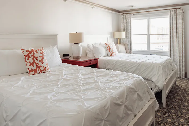 Modern hotel room with two white queen beds, coral patterned pillows, and patterned carpet flooring.