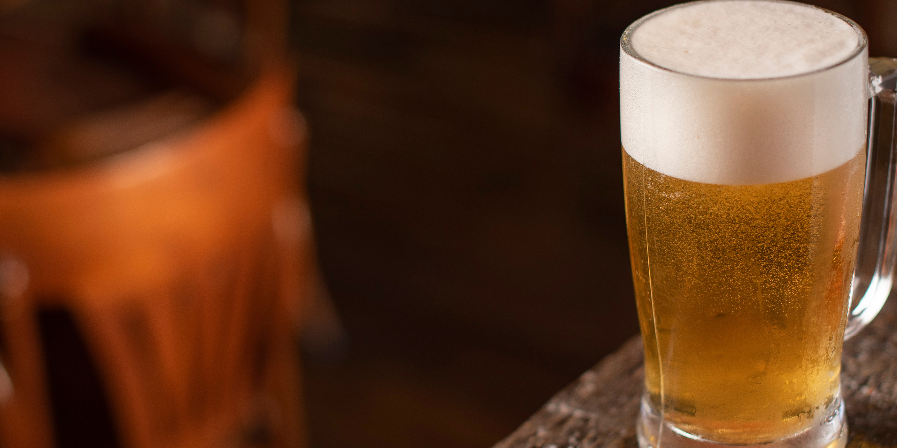 Cold beer in a glass mug with frothy foam sitting on a wooden bar top.