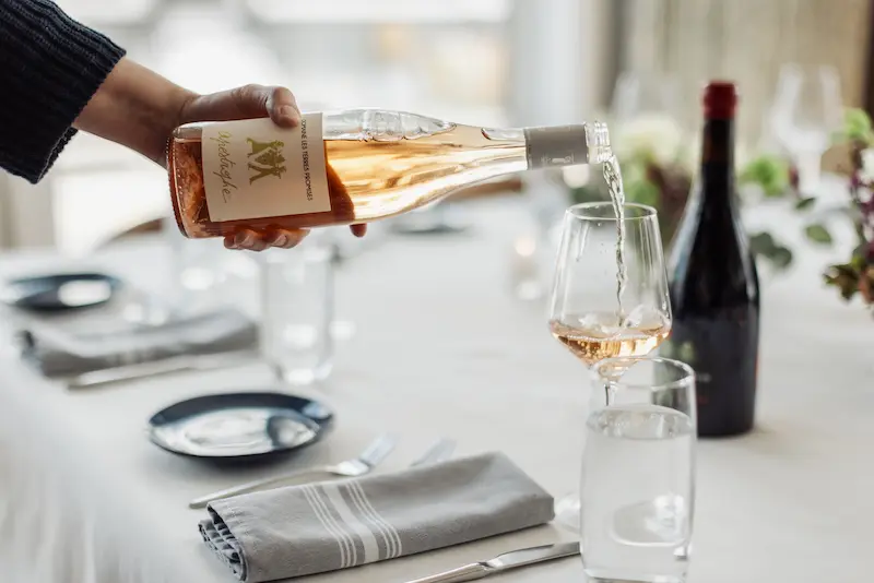 A hand pours Domaine Les Terres Promises rosé wine into a glass at a formal dining table setting.