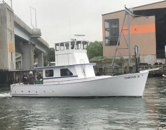 The Tartan II white fishing boat cruises on the water near a bridge and industrial building.