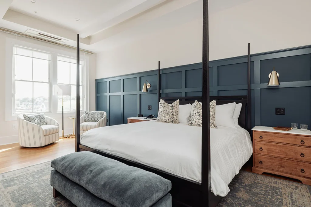 Modern hotel bedroom featuring a four-poster bed, deep blue paneled accent wall, and cozy seating area by the window.