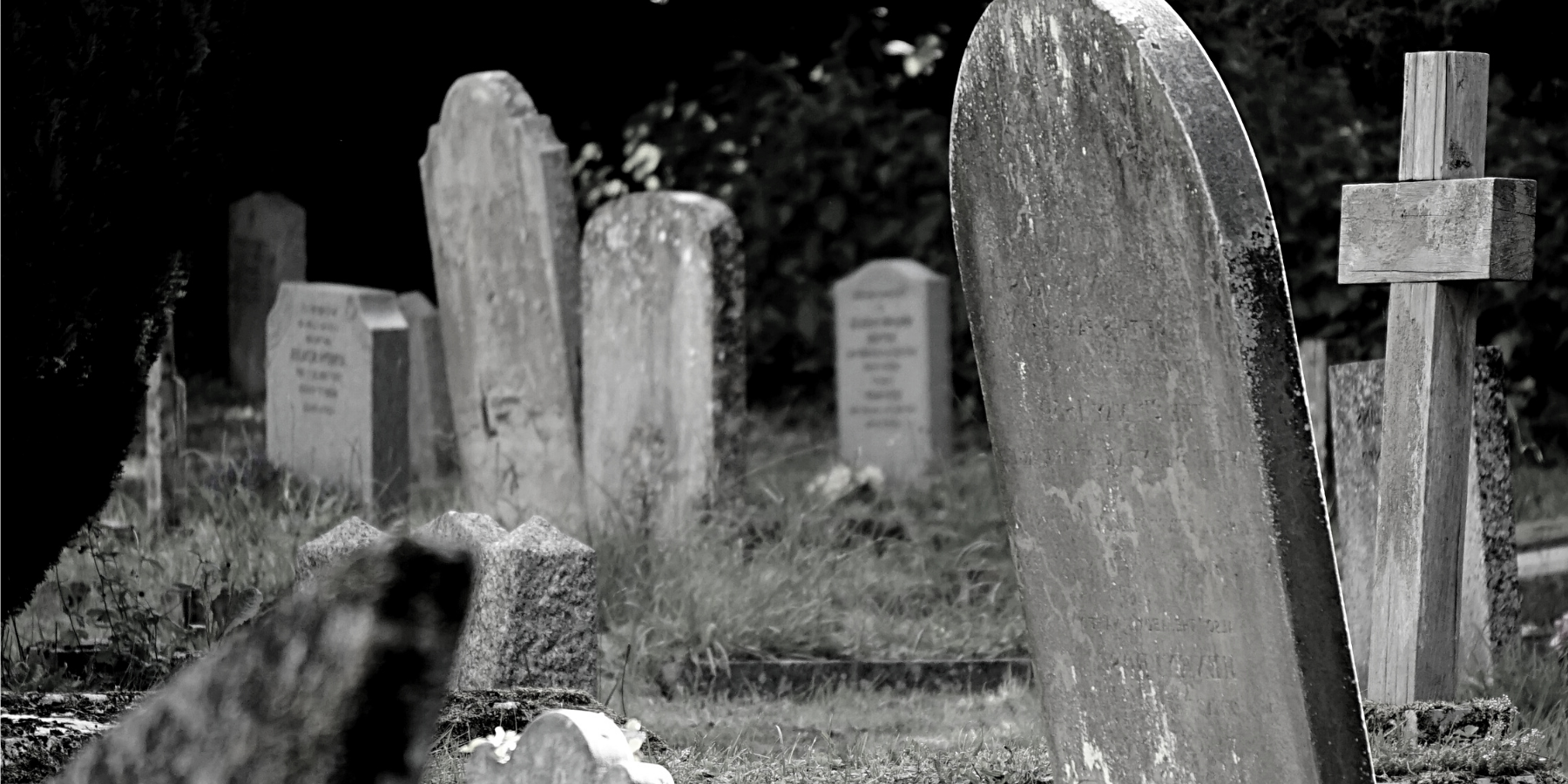 Black and white photograph of an old, weathered graveyard with several stone headstones and a wooden cross.