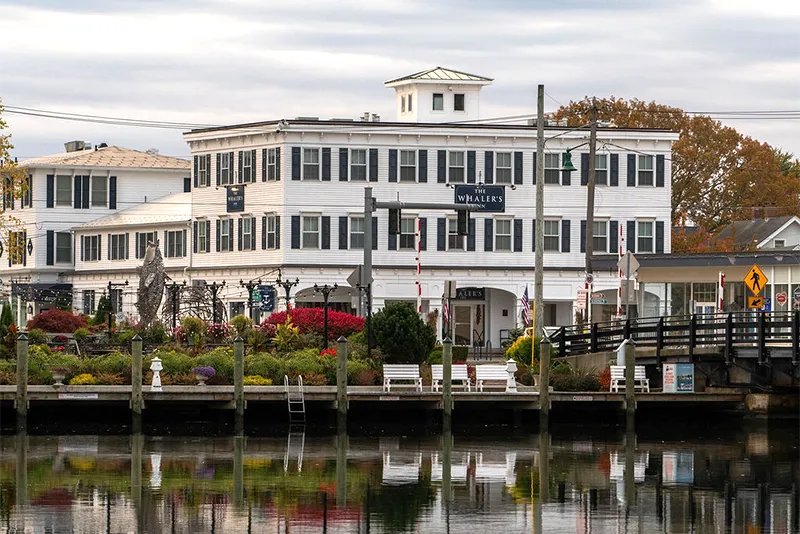 A white, multi-story waterfront hotel with dark shutters, situated behind a dock with a reflection in the water.