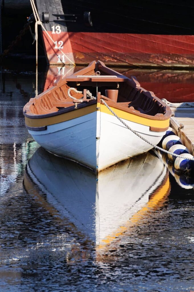 A white and yellow wooden rowboat floats in calm harbor waters with a clear reflection near a larger vessel.