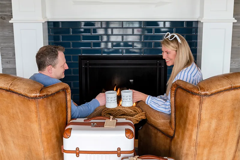 Couple relaxing with coffee mugs by a cozy fireplace next to vintage travel suitcases at a boutique hotel.