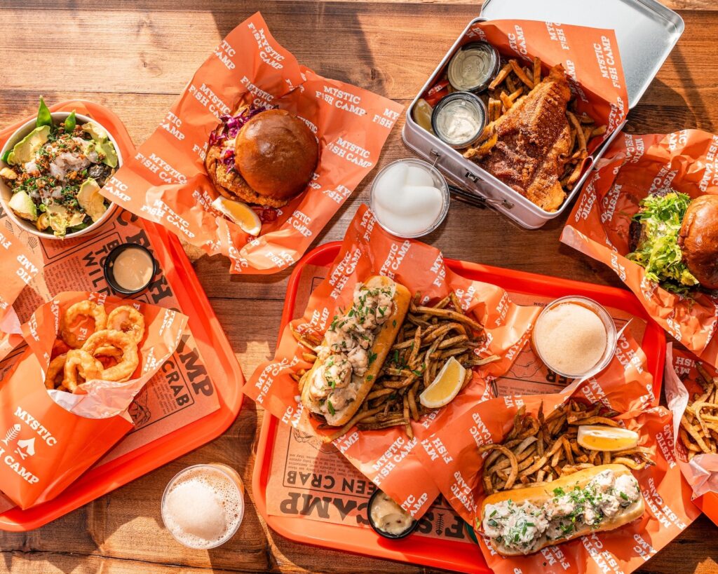 Top-down view of Mystic Fish Camp seafood, including lobster rolls, fried fish, burgers, and sides on orange paper.