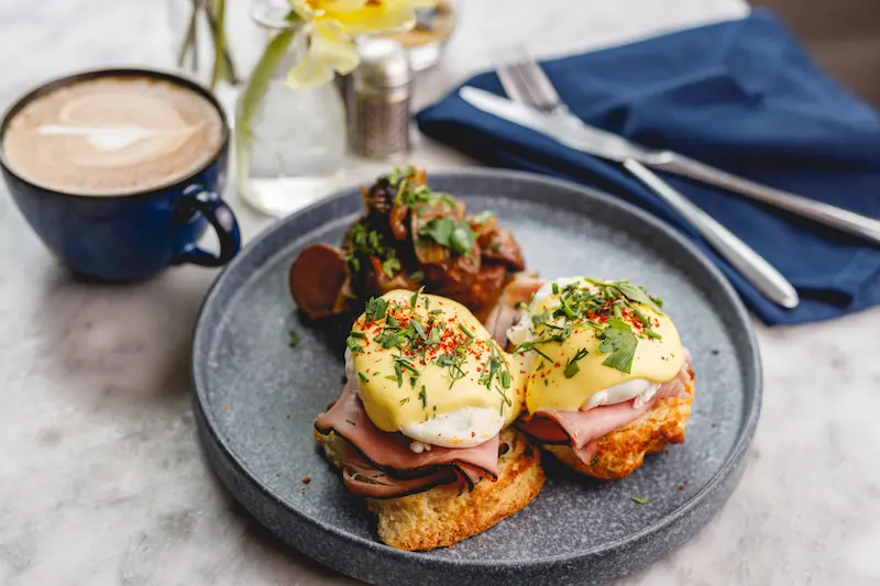 Classic Eggs Benedict with ham and hollandaise sauce served on a plate with mushrooms and a cup of coffee.