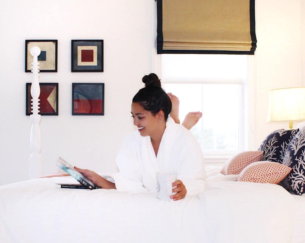 Woman in a white bathrobe relaxing on a bed, reading a book, and holding a mug in a bright, modern bedroom.