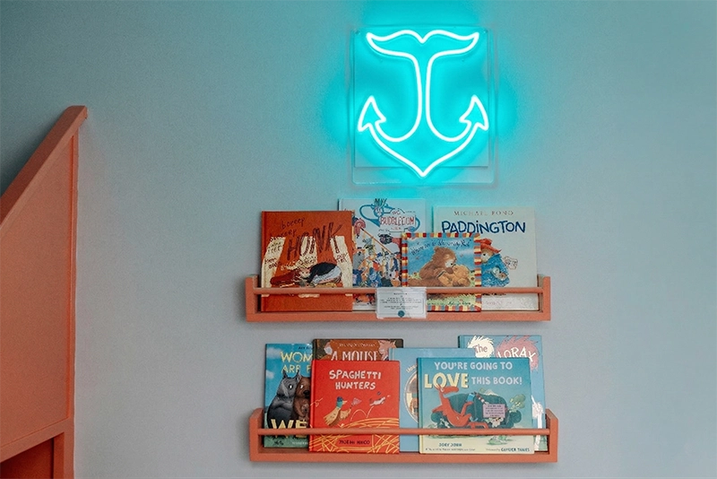 Floating orange nursery bookshelves display a collection of colorful children's books under a glowing neon anchor sign.