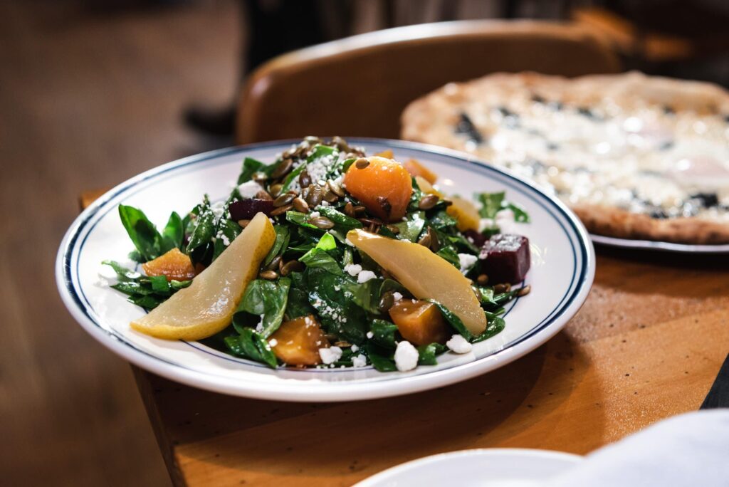 Fresh spinach salad with roasted beets, sliced pears, pumpkin seeds, and goat cheese on a restaurant plate.