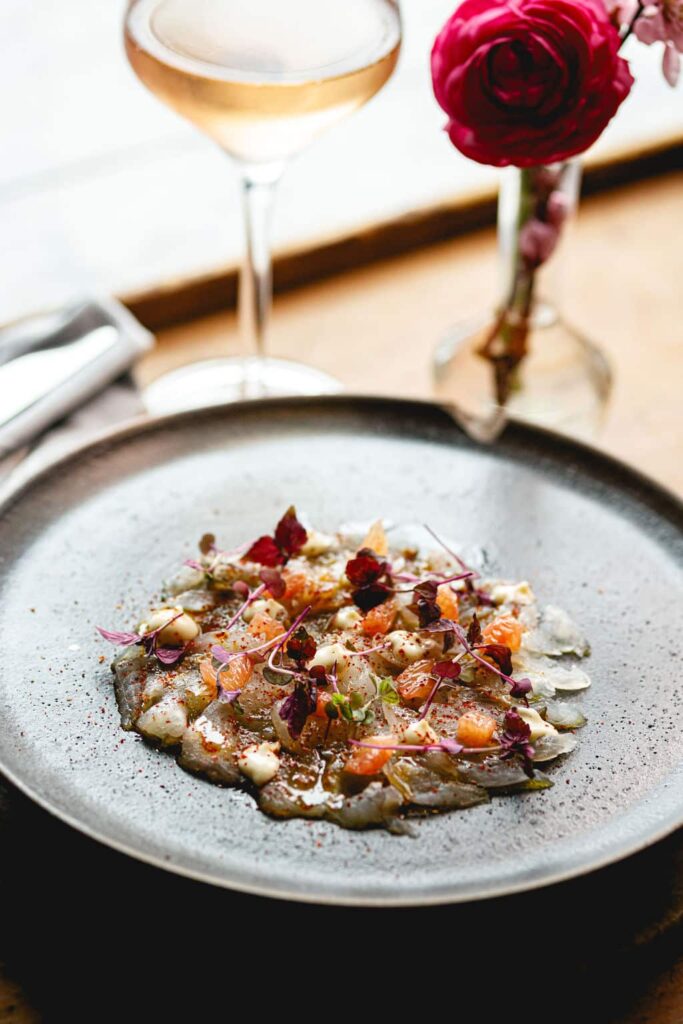 Exquisite raw fish carpaccio garnished with microgreens and citrus on a grey ceramic plate with a glass of rosé.