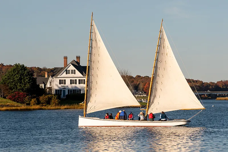 A wooden sailboat with white sails cruises on a calm river past a large white colonial-style home.