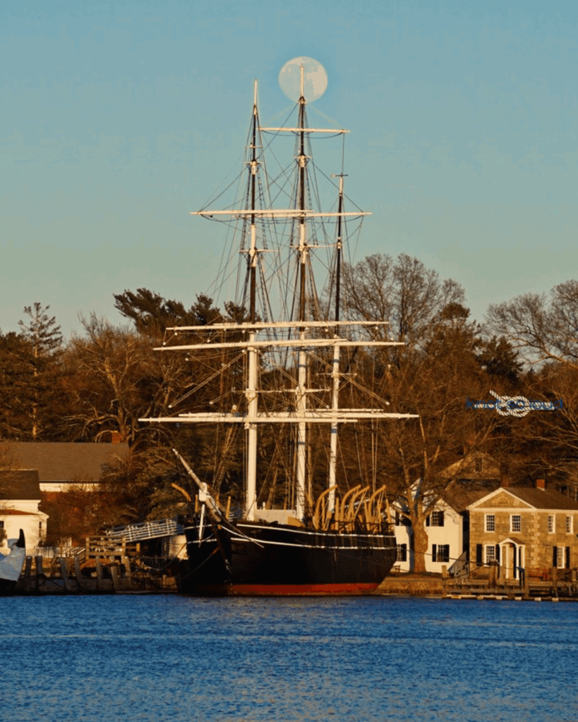 A historic tall ship docked at Mystic Seaport with a full moon positioned perfectly behind its main mast.