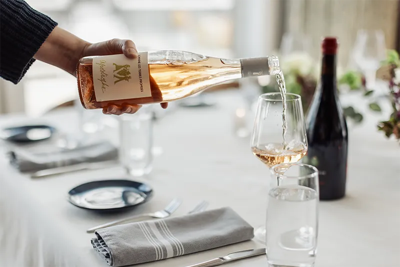 A hand pours Domaine Les Terres Promises rosé wine into a glass on an elegantly set restaurant dining table.