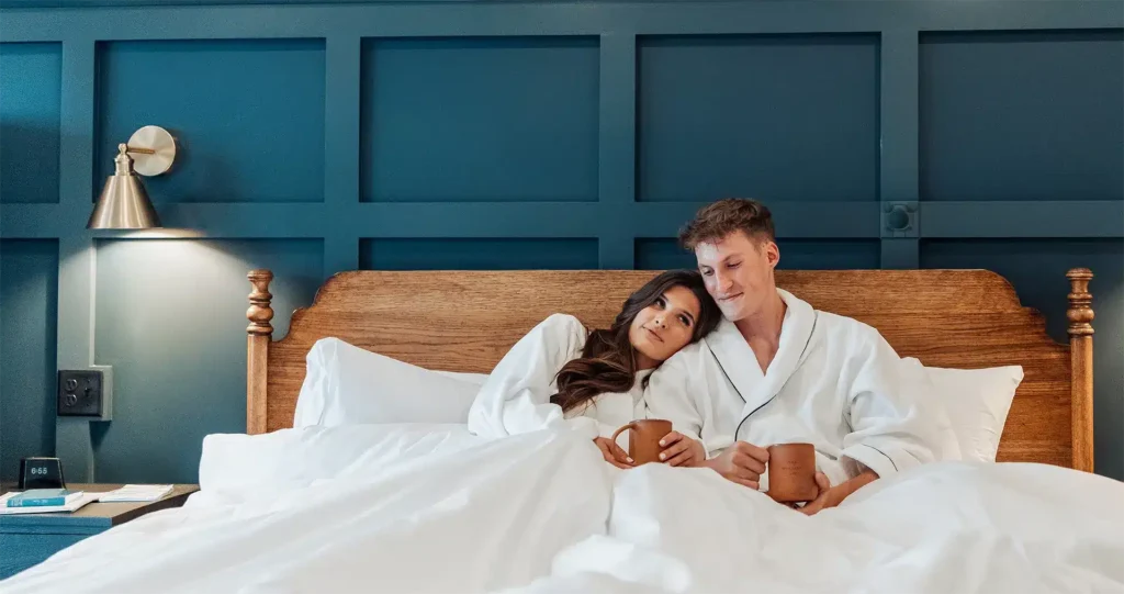 A couple in white robes sits in a hotel bed with coffee mugs against a dark blue paneled wall.