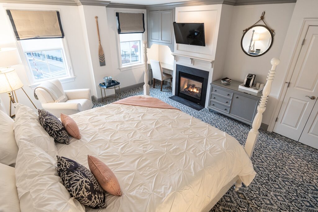 Cozy hotel bedroom with a fireplace, king bed, patterned carpet, and coastal decor.