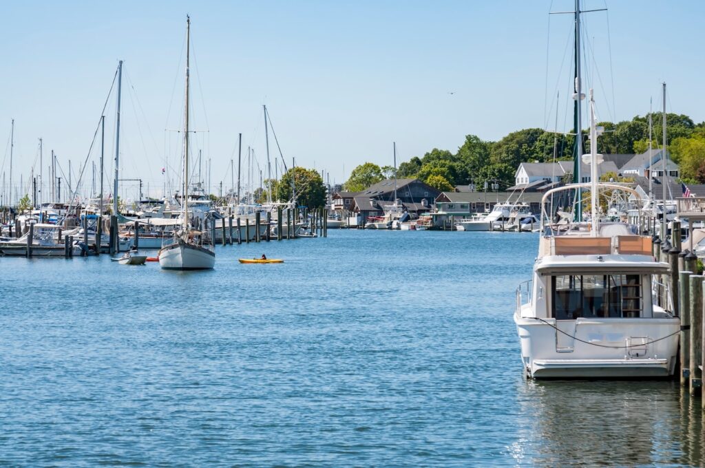 A sunny harbor scene featuring docked sailboats and a motor yacht with a kayaker paddling through calm waters.