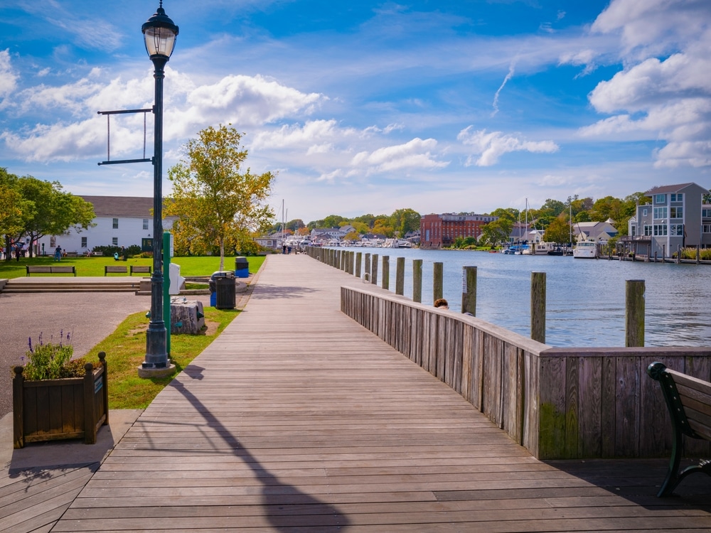 Wooden boardwalk along the harbor in Mystic, Connecticut, with boats and scenic waterfront buildings under a blue sky.