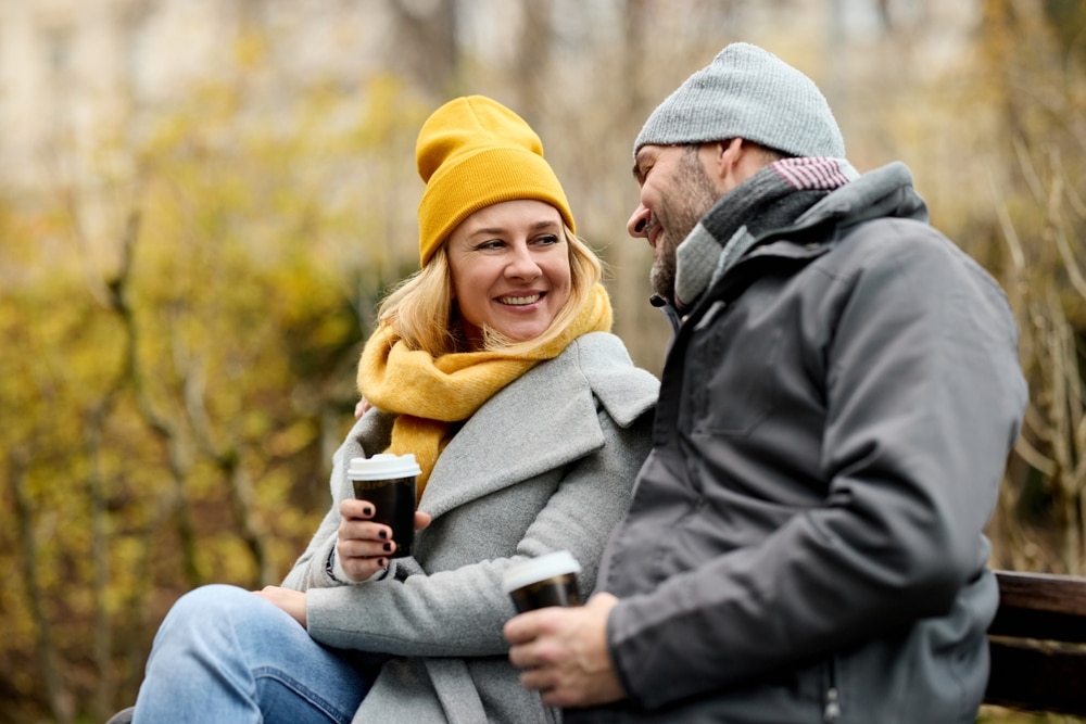 Happy couple sitting on a park bench in autumn wearing warm clothing and drinking coffee.