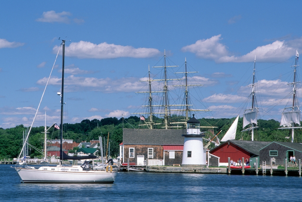 A sailboat sails past the Mystic Seaport lighthouse and historic tall ships on a sunny summer day.