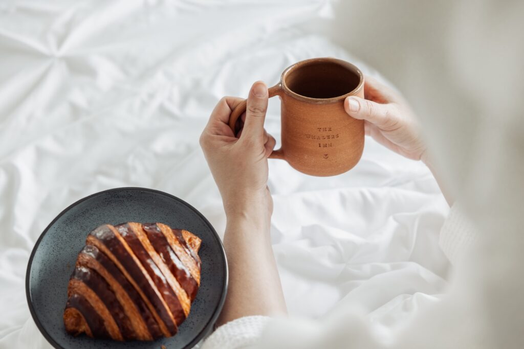 A chocolate croissant on a bed framed by a person holding a coffee cup that says, "The Whaler's Inn," one of the best boutique hotels in Mystic CT.